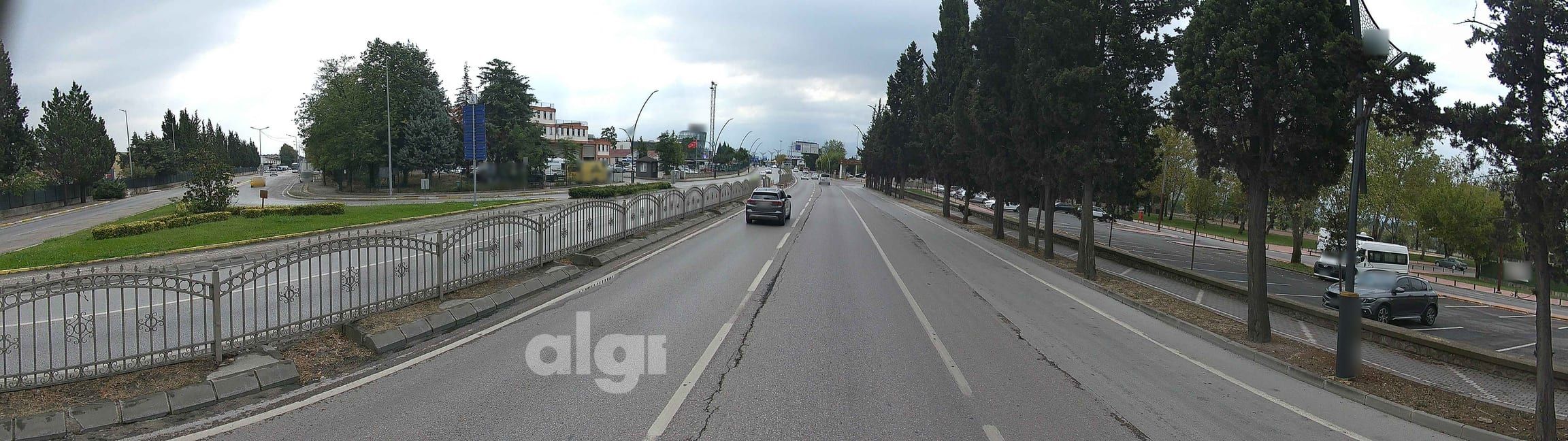 Araç kamerasından iki yönlü bir cadde: ortada korkuluklu refüj, solda şerit çizgili yol yüzeyi, sağda ağaçlı kaldırım ve park alanı